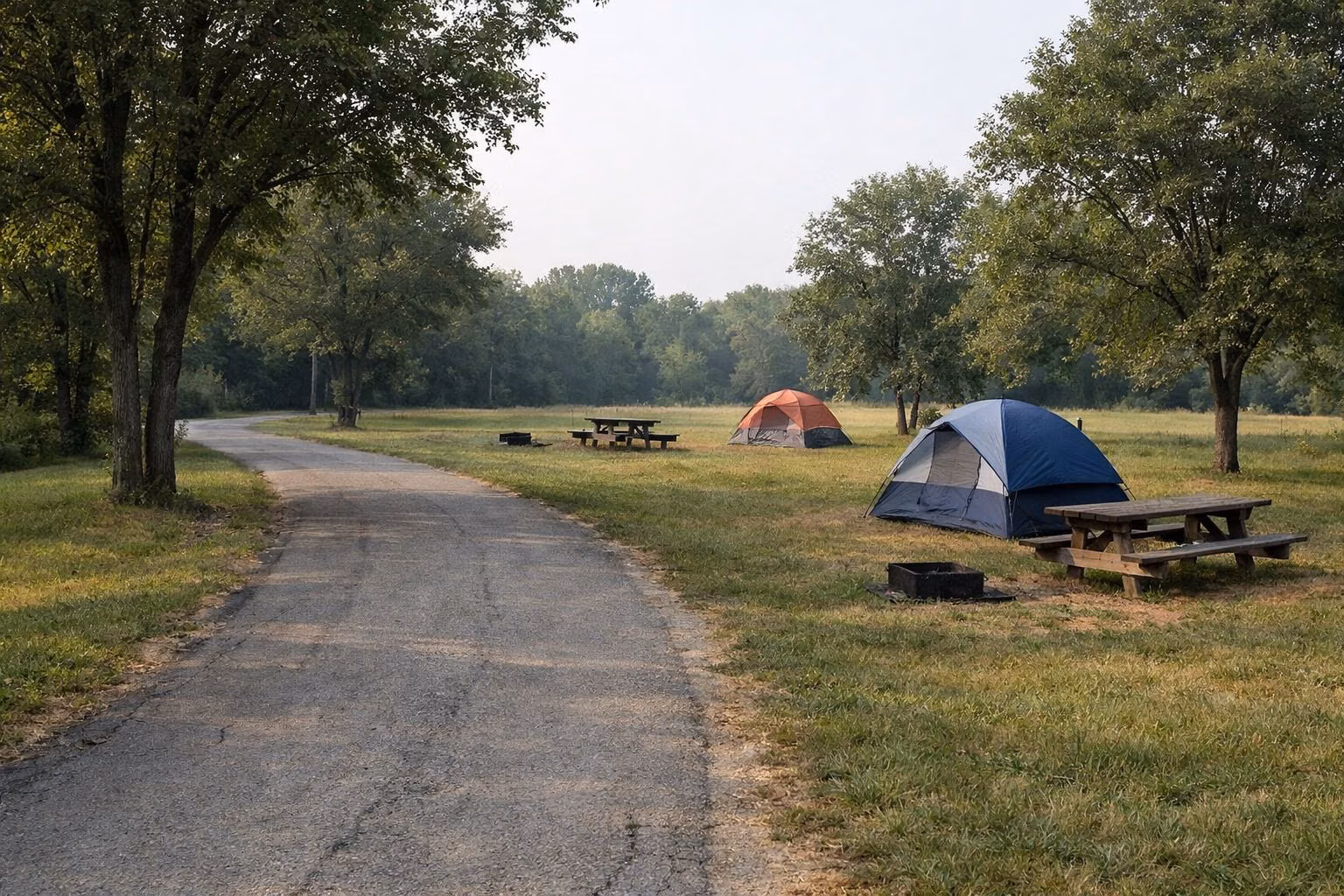 Worn paved campground road past grassy campsites with tents, picnic tables and fire pits