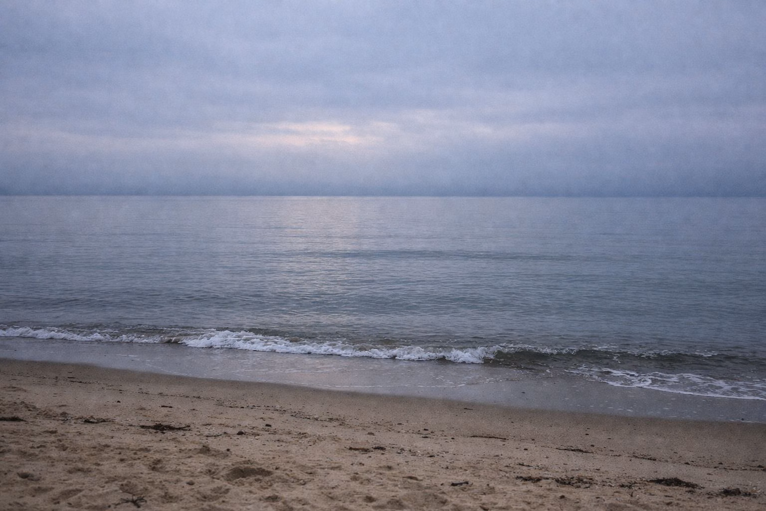Gray dawn light over calm water seen from a sandy beach, with small waves at the edge