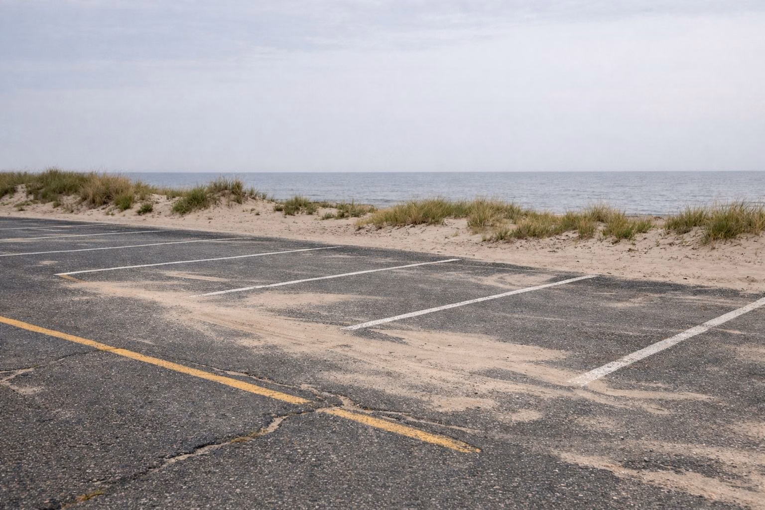 Empty beach parking lot with faded lines and blown sand, low dunes and water beyond