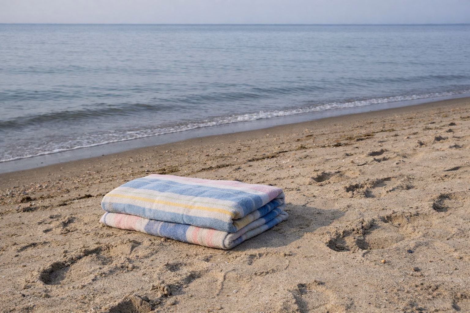 Folded striped beach towel on the sand near calm water, with footprints around it