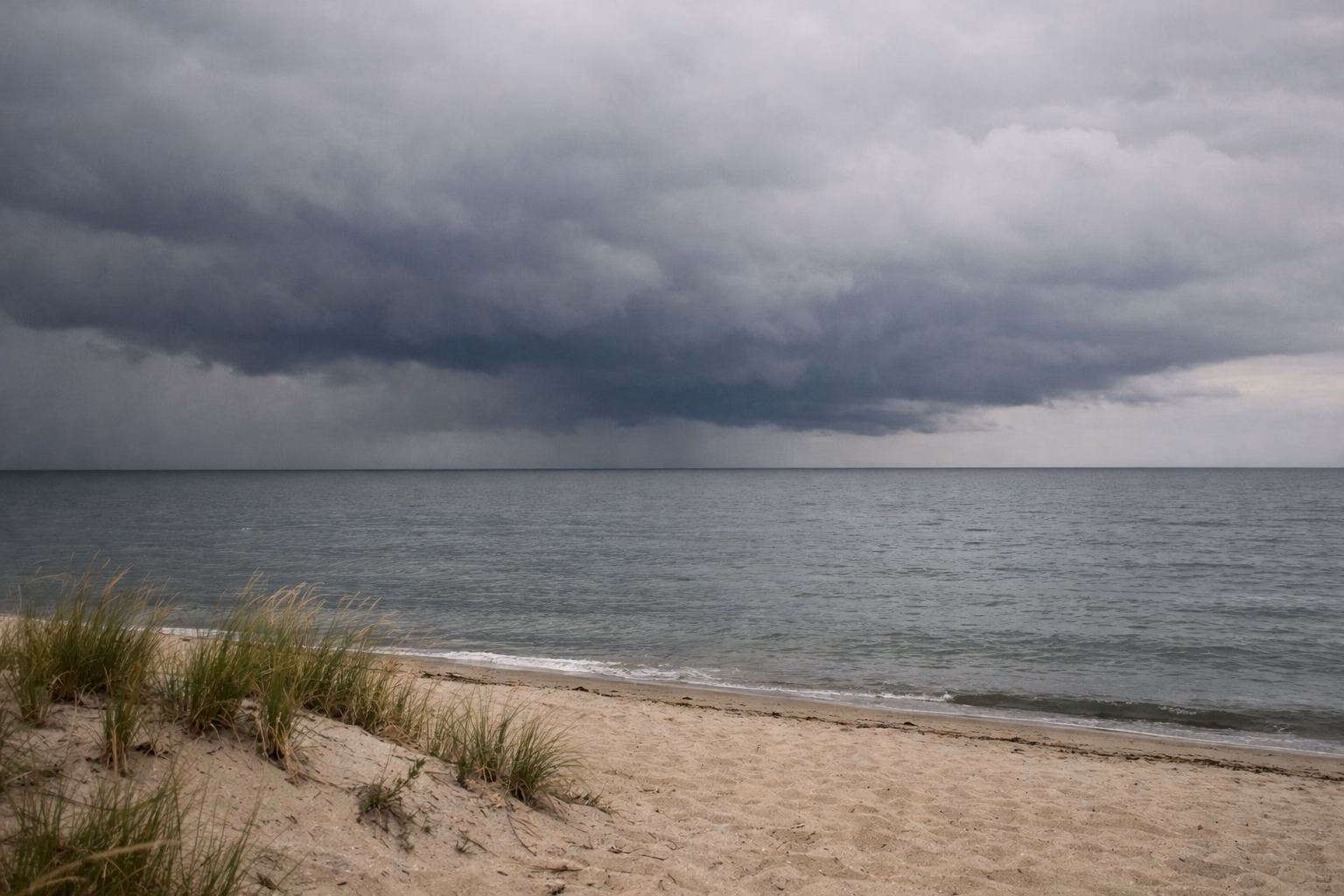 Dark storm clouds building over calm gray water beyond an empty sandy beach with dune grass